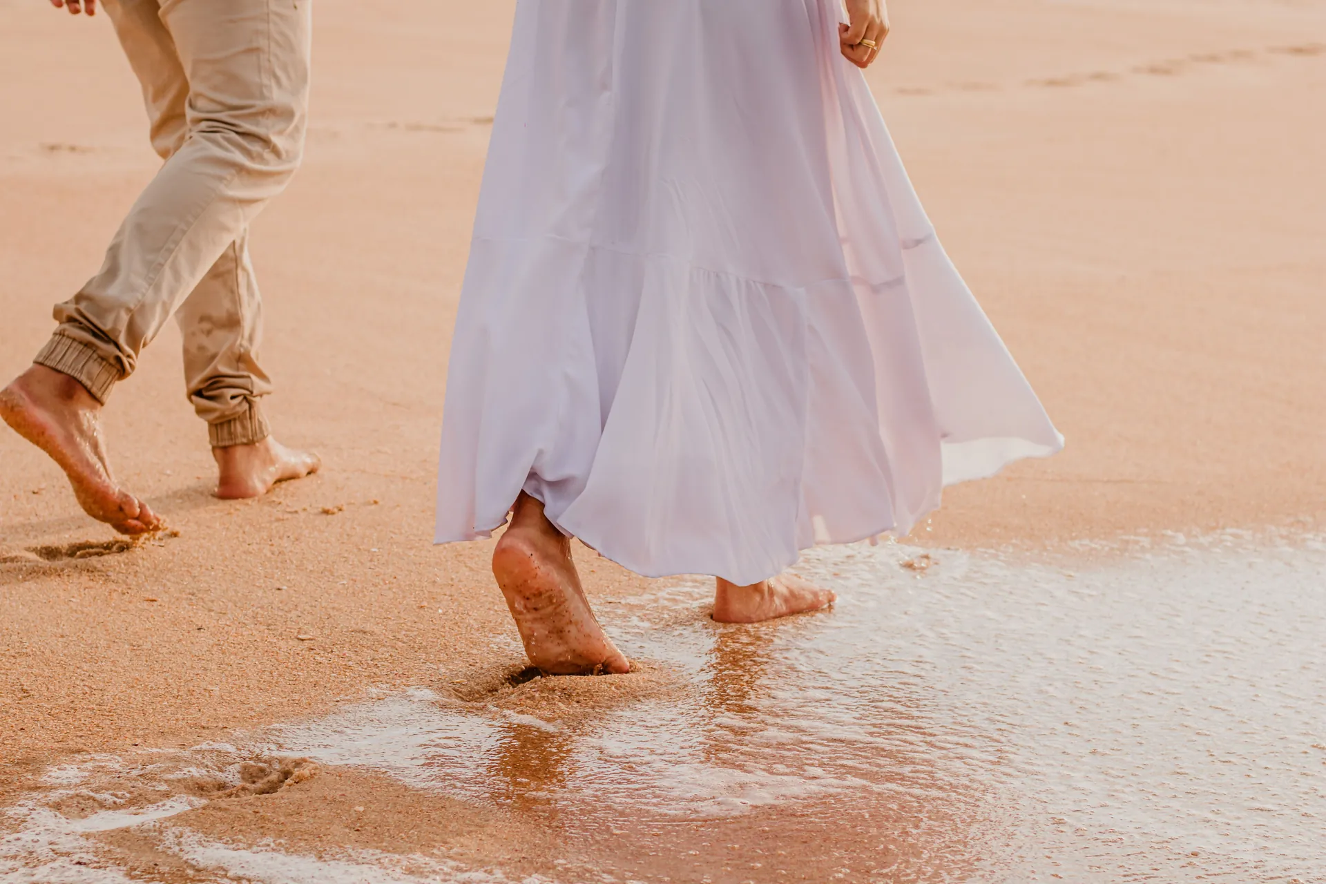 Pés descalços de um casal na beira d’água, com o vestido branco dela ao vento