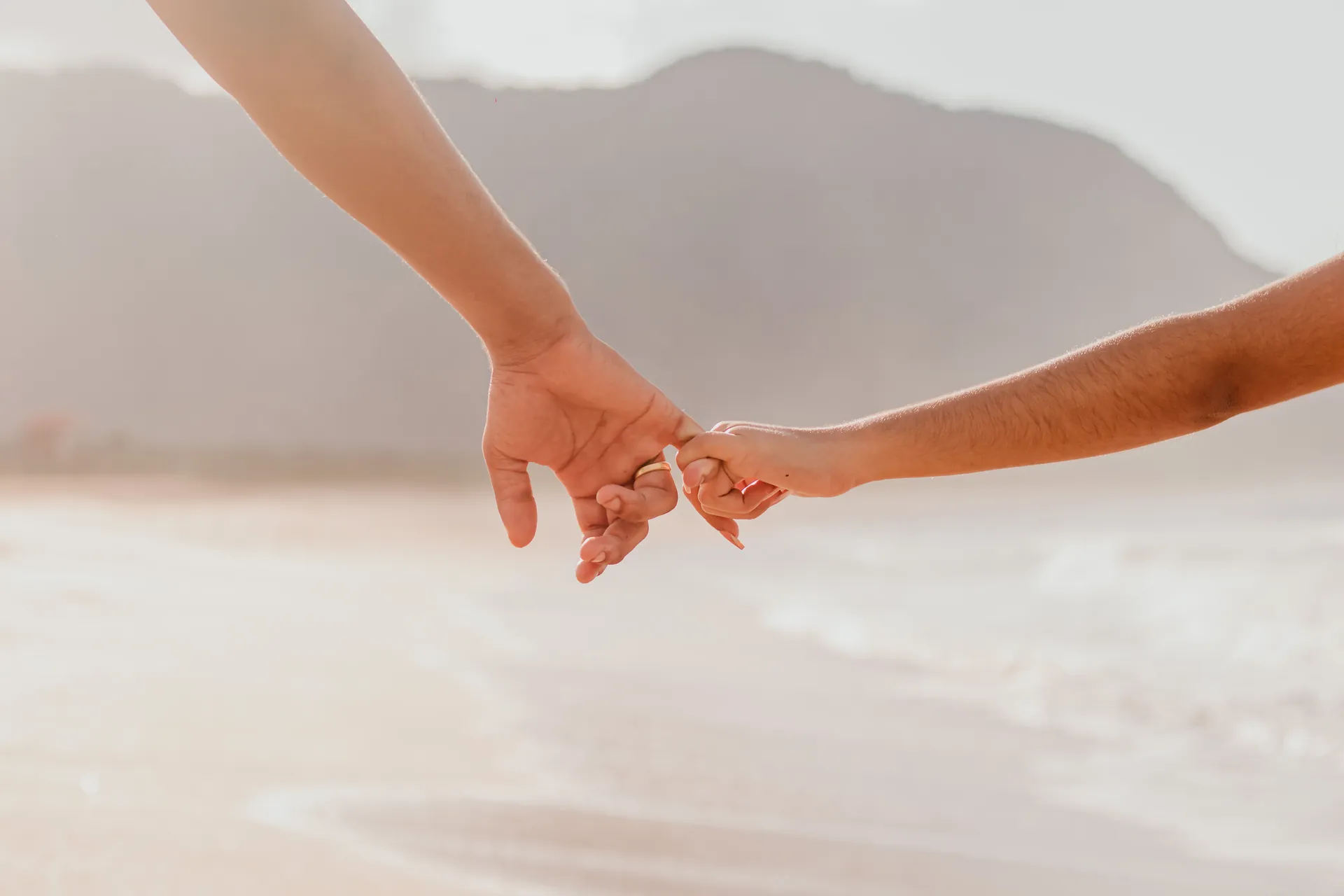 Mãos de um casal se encontrando, com a praia clara desfocada ao fundo