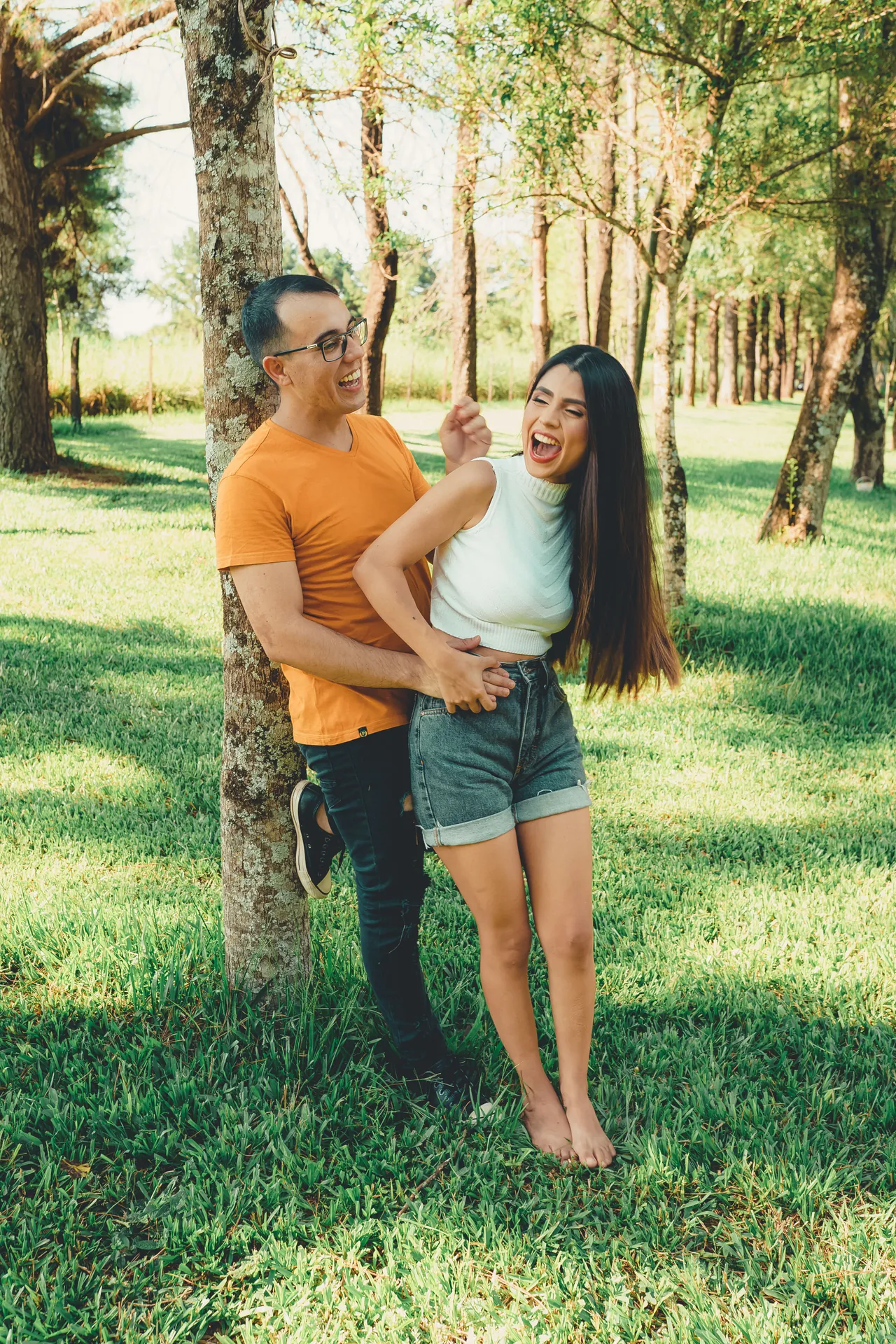 Casal rindo abraçado, encostado numa árvore de parque