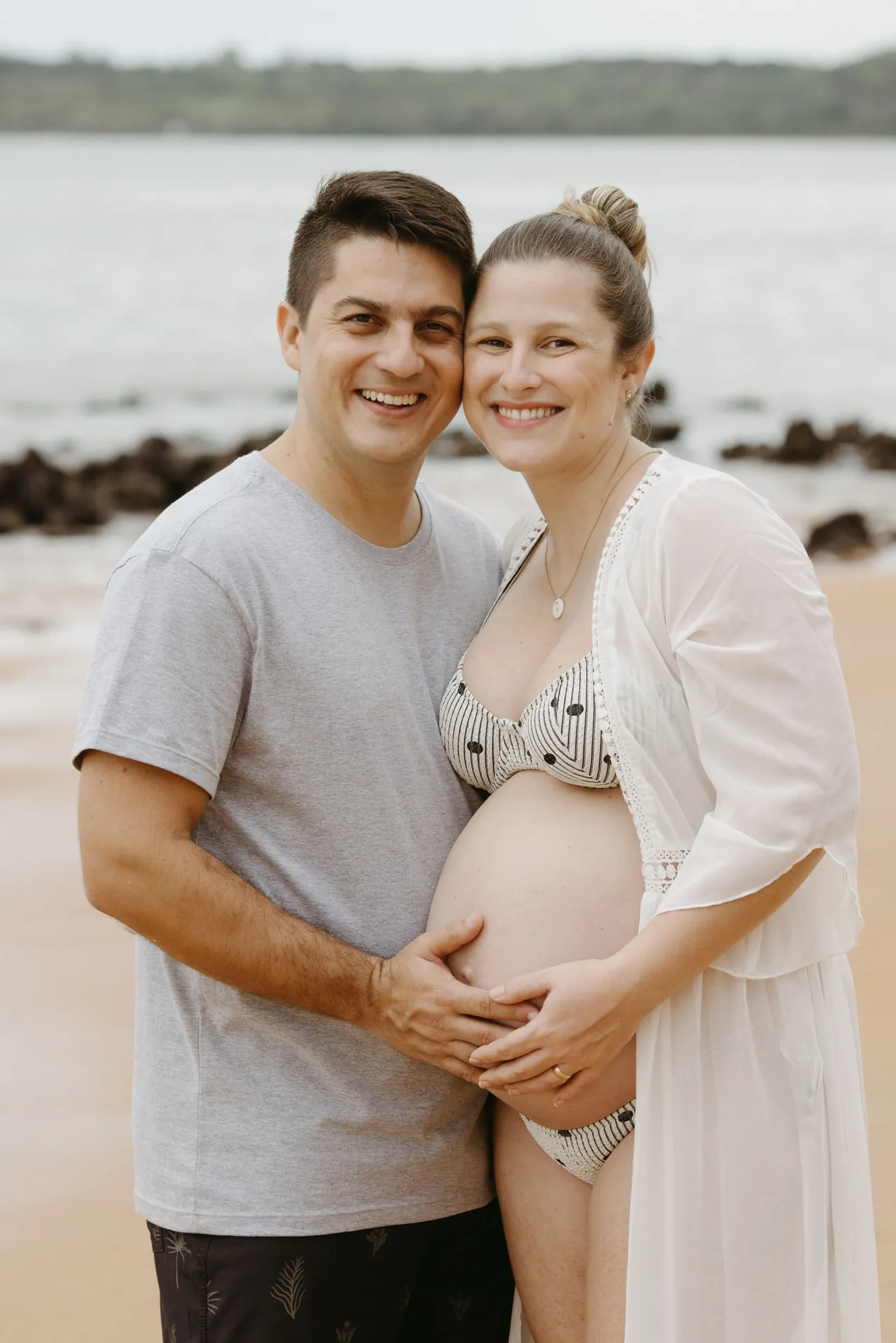 Casal abraçado com as mãos sobre a barriga da gestante, na praia