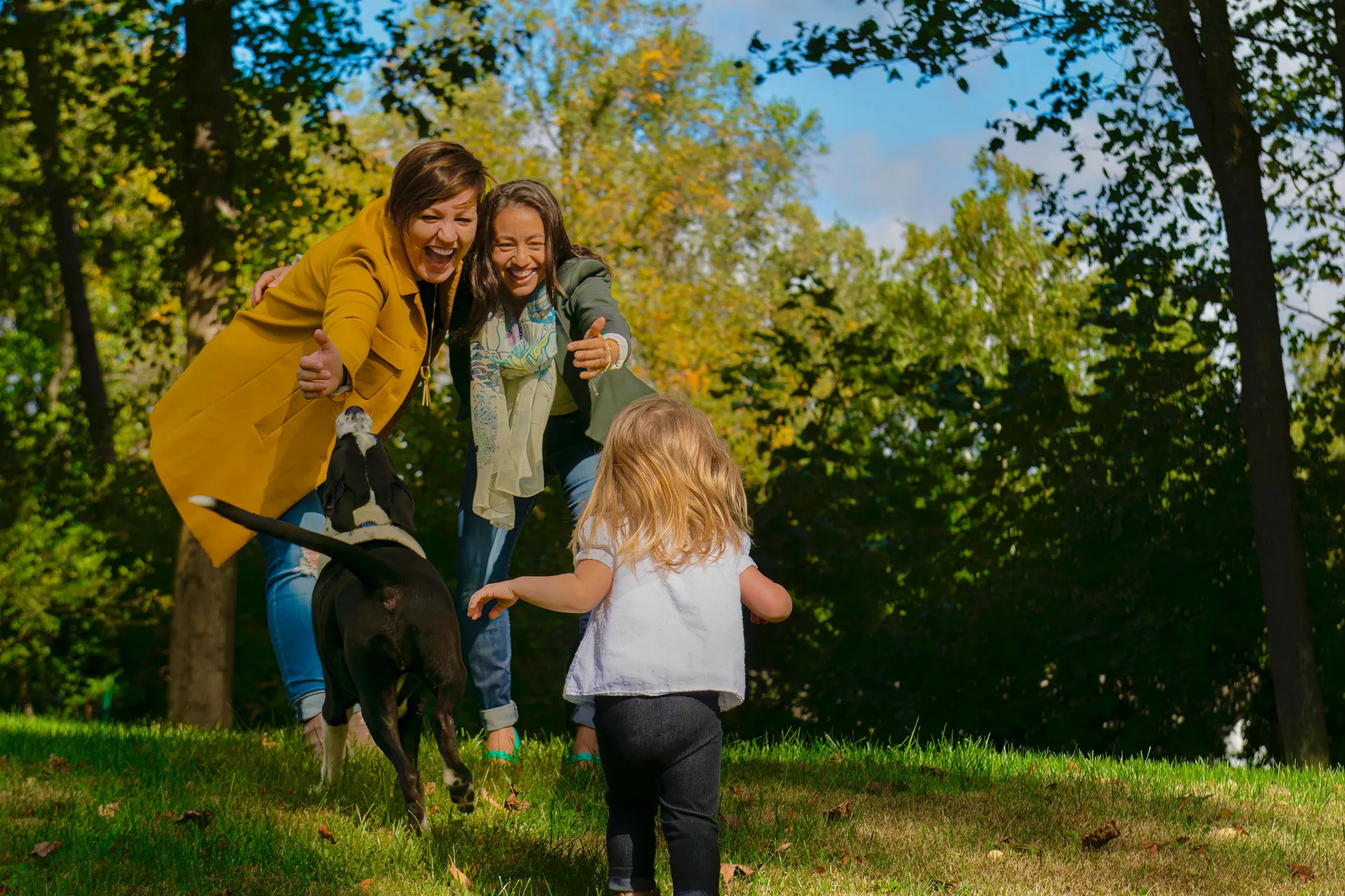 Criança pequena correndo pros braços de duas mulheres que riem, com o cachorro da família ao lado, num parque