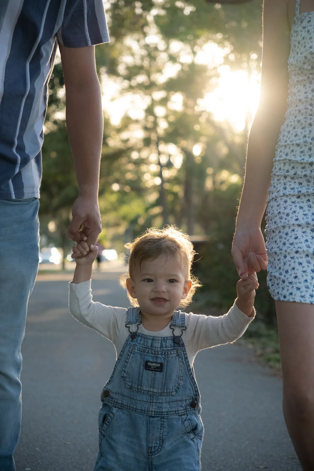 Criança pequena de jardineira andando de mãos dadas com os pais, em contraluz dourado