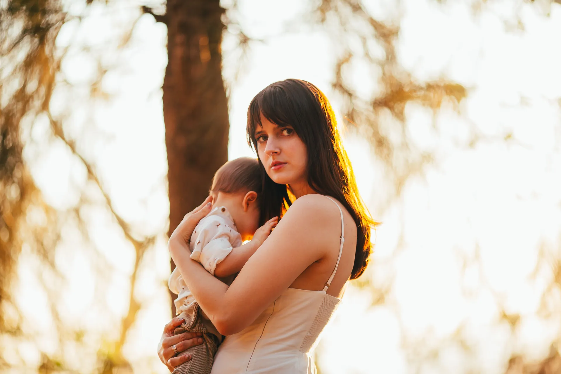 Mãe com o bebê adormecido no colo, entre árvores na luz dourada do fim de tarde