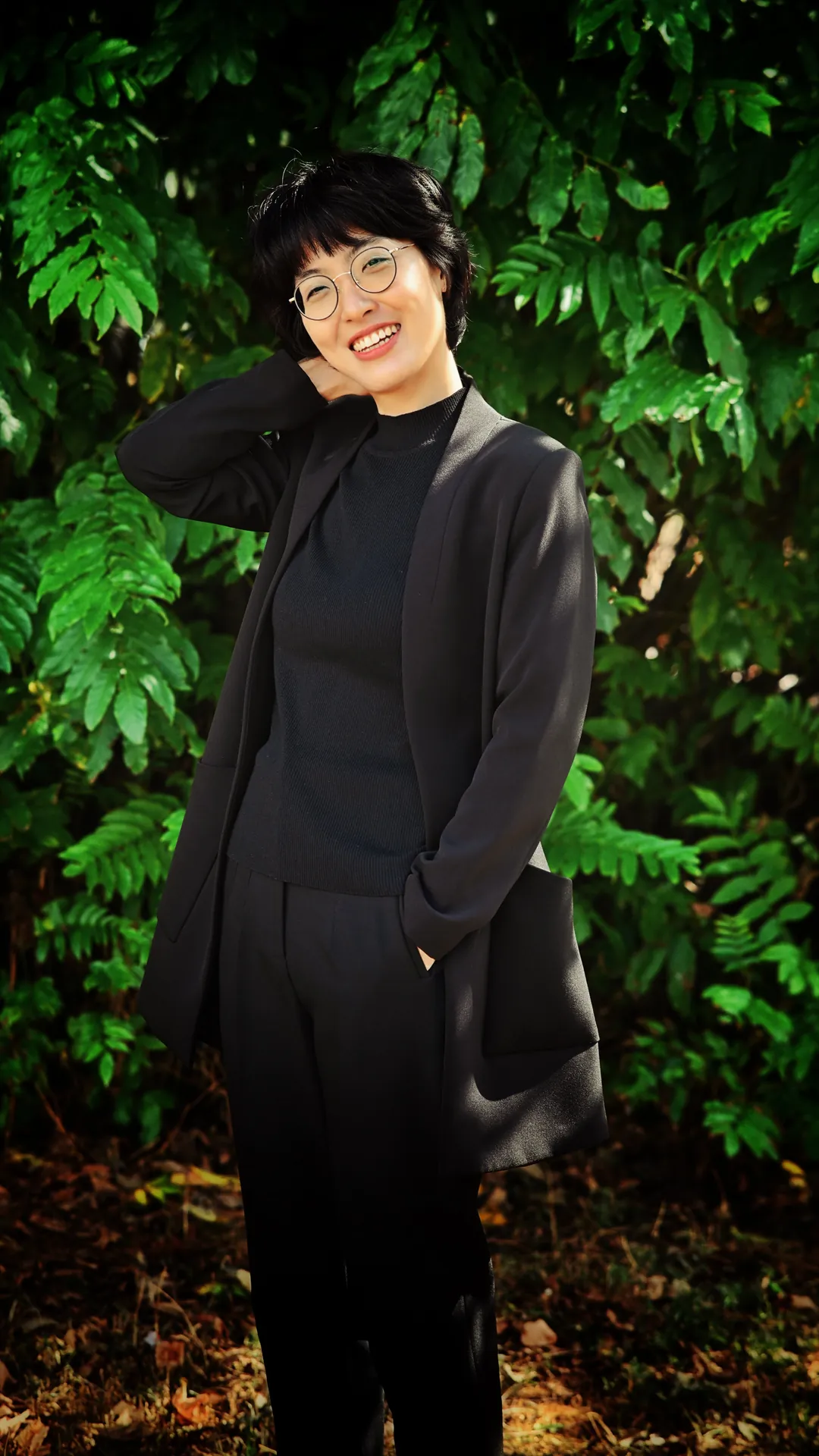 Mulher de blazer escuro e óculos sorrindo diante de folhagens
