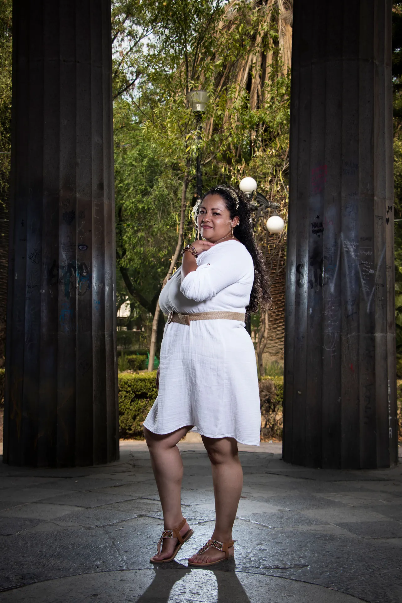 Mulher plus size de vestido branco posando entre colunas num parque, na luz dourada