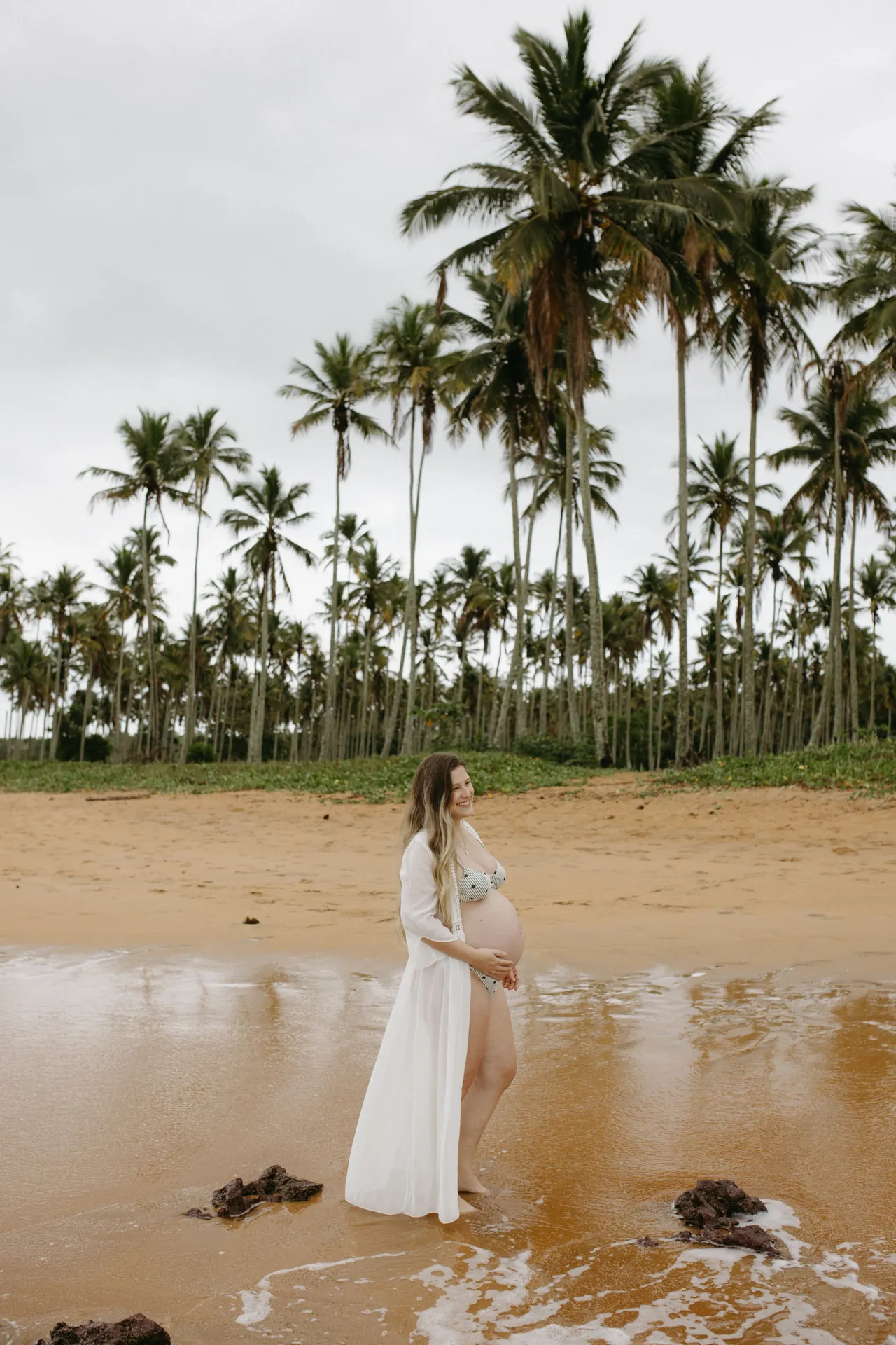 Gestante de vestido branco na beira d’água, com coqueiros ao fundo