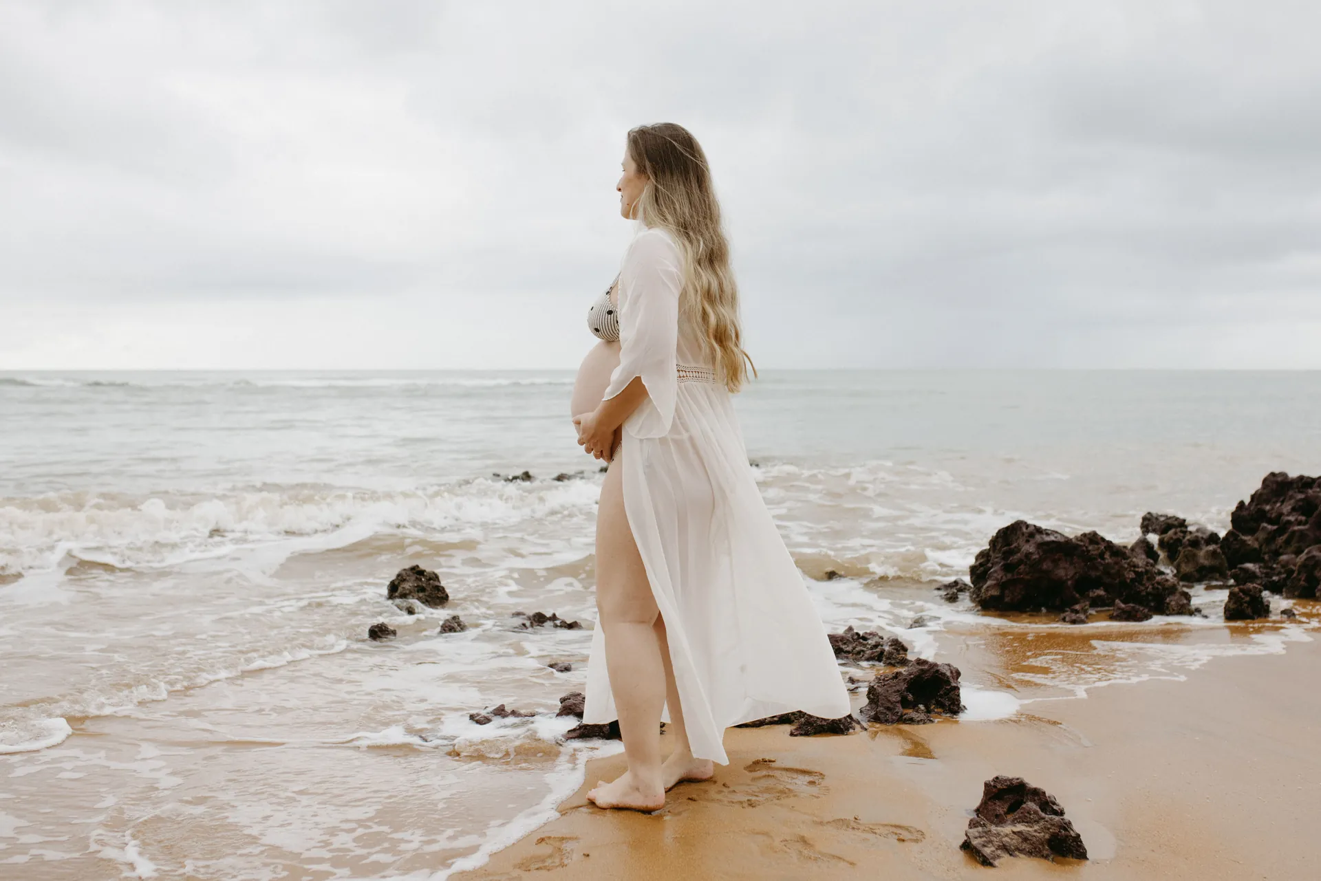 Gestante de vestido branco à beira do mar, olhando o horizonte