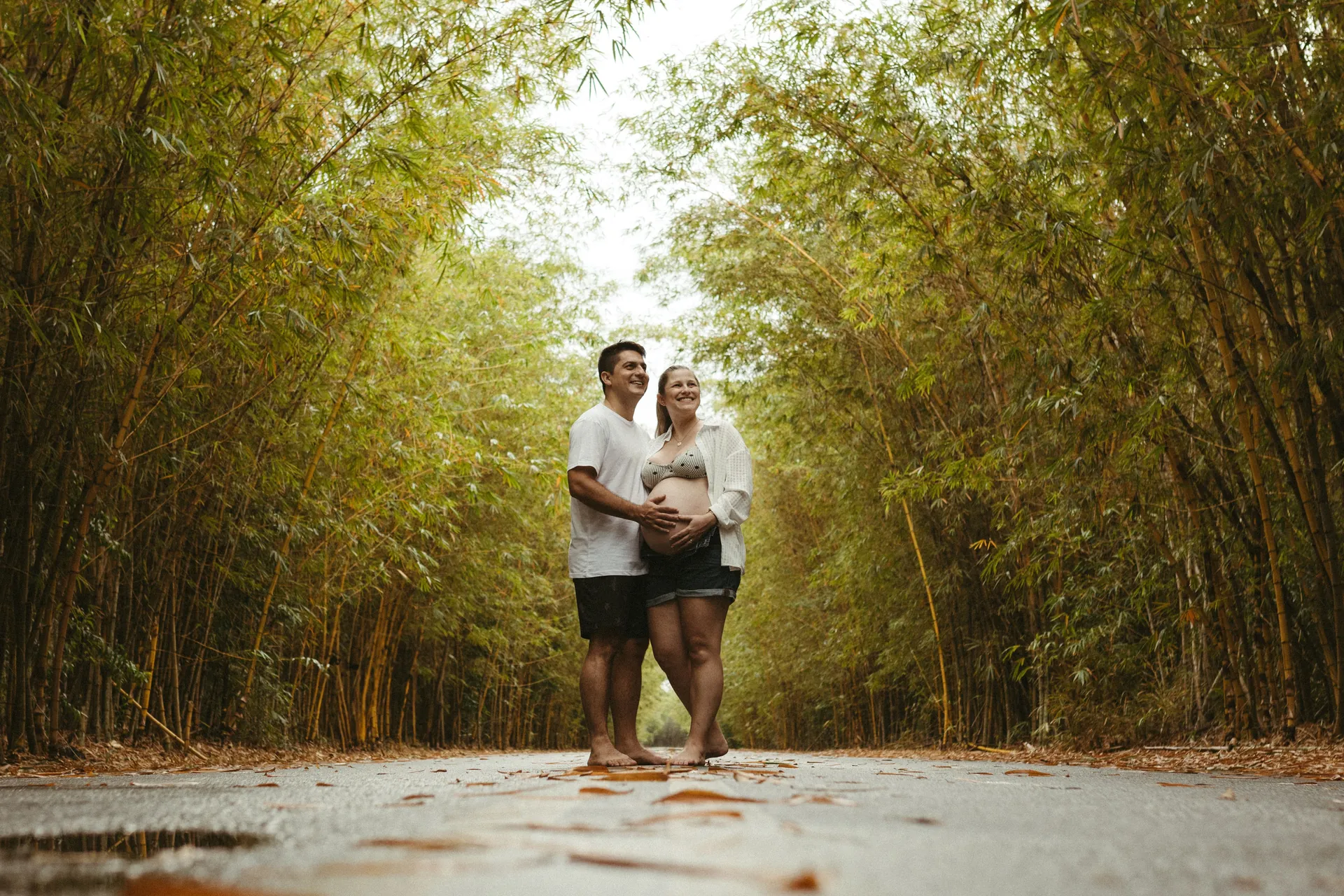 Casal grávido abraçado no meio de uma estrada cercada de bambus