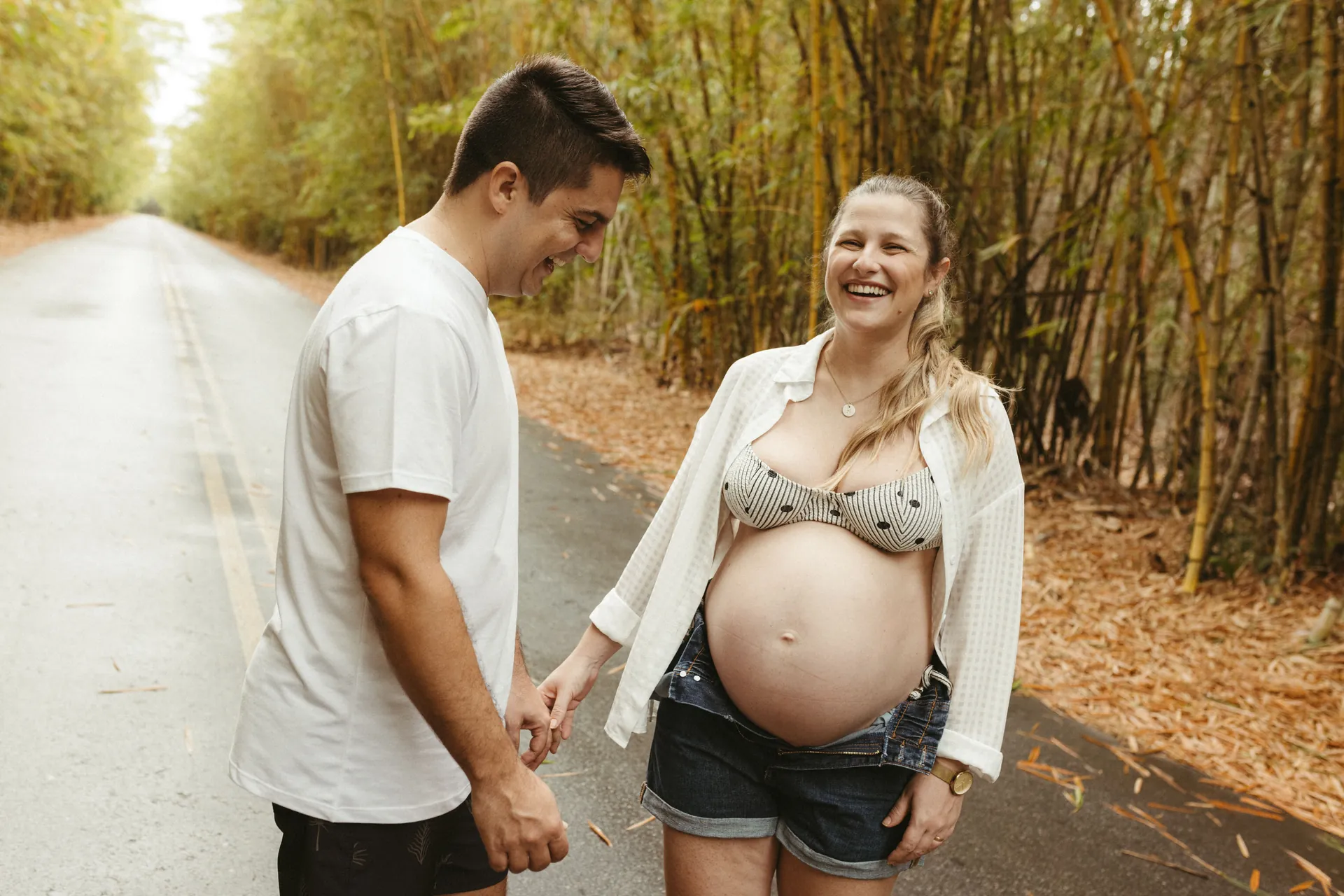 Casal grávido rindo de mãos dadas ao ar livre, ela com a barriga à mostra