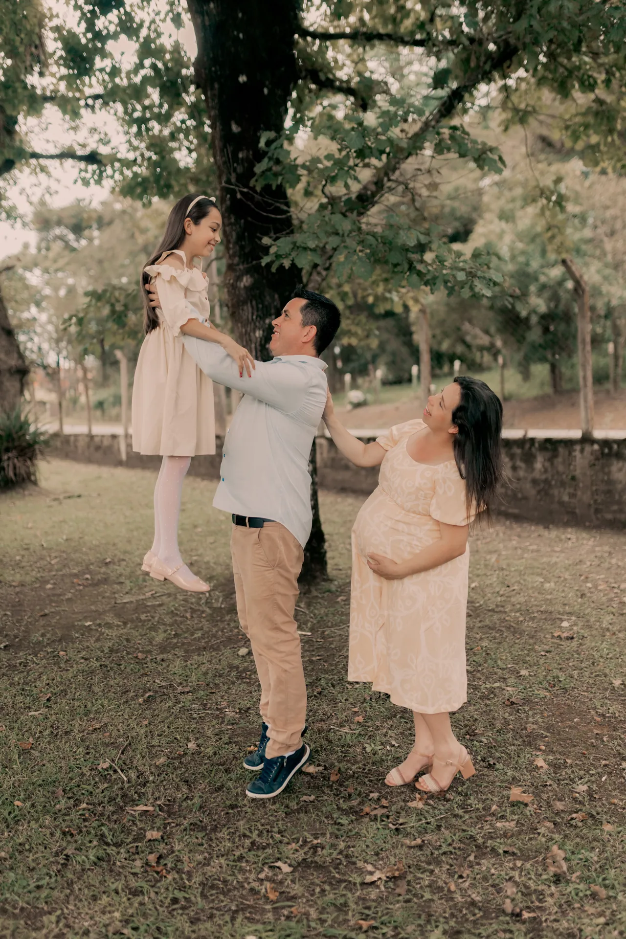 Pai erguendo a filha no alto enquanto a mãe grávida observa sorrindo, num parque