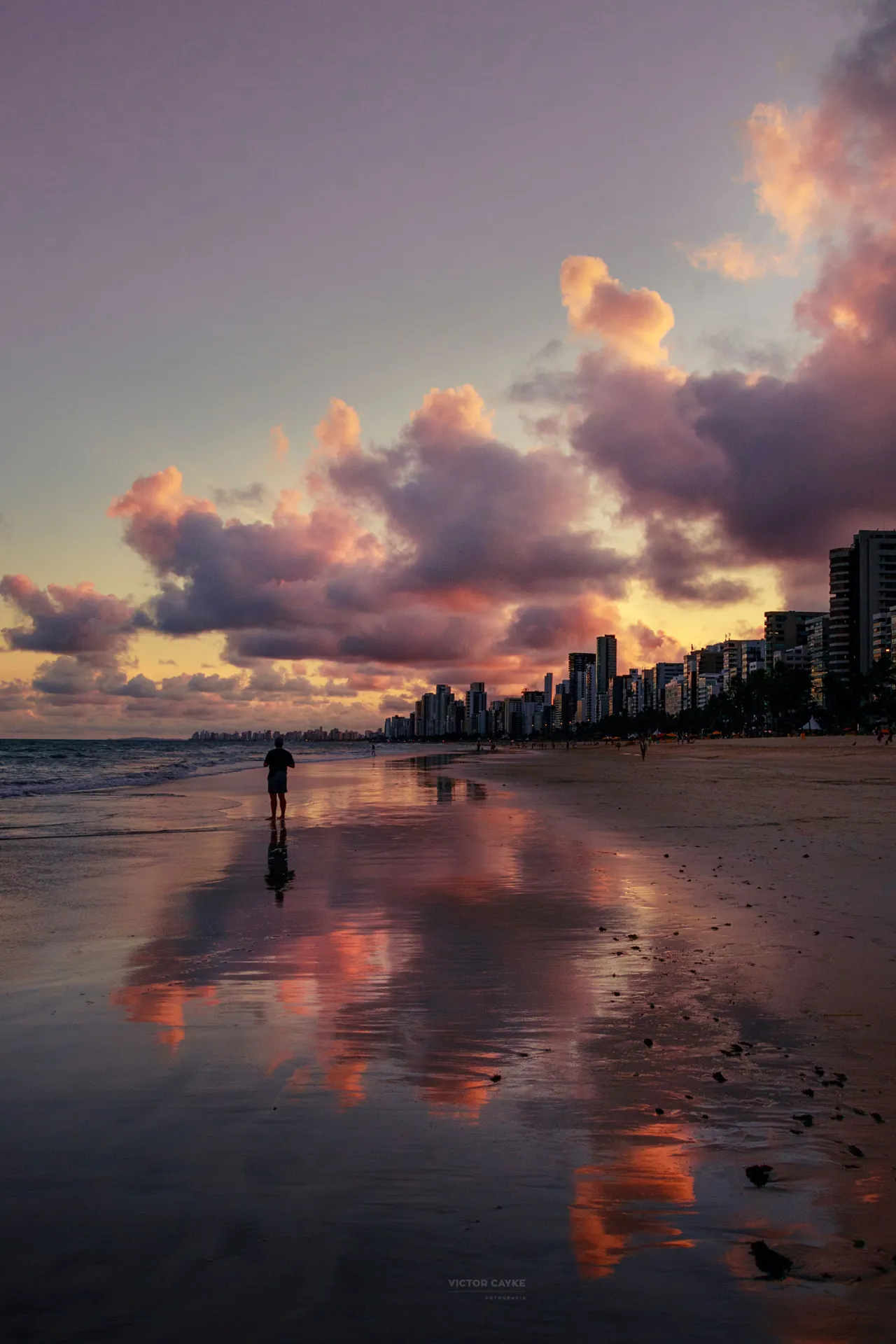 Praia de Boa Viagem no fim de tarde, com a areia molhada refletindo as nuvens rosadas e os prédios da orla