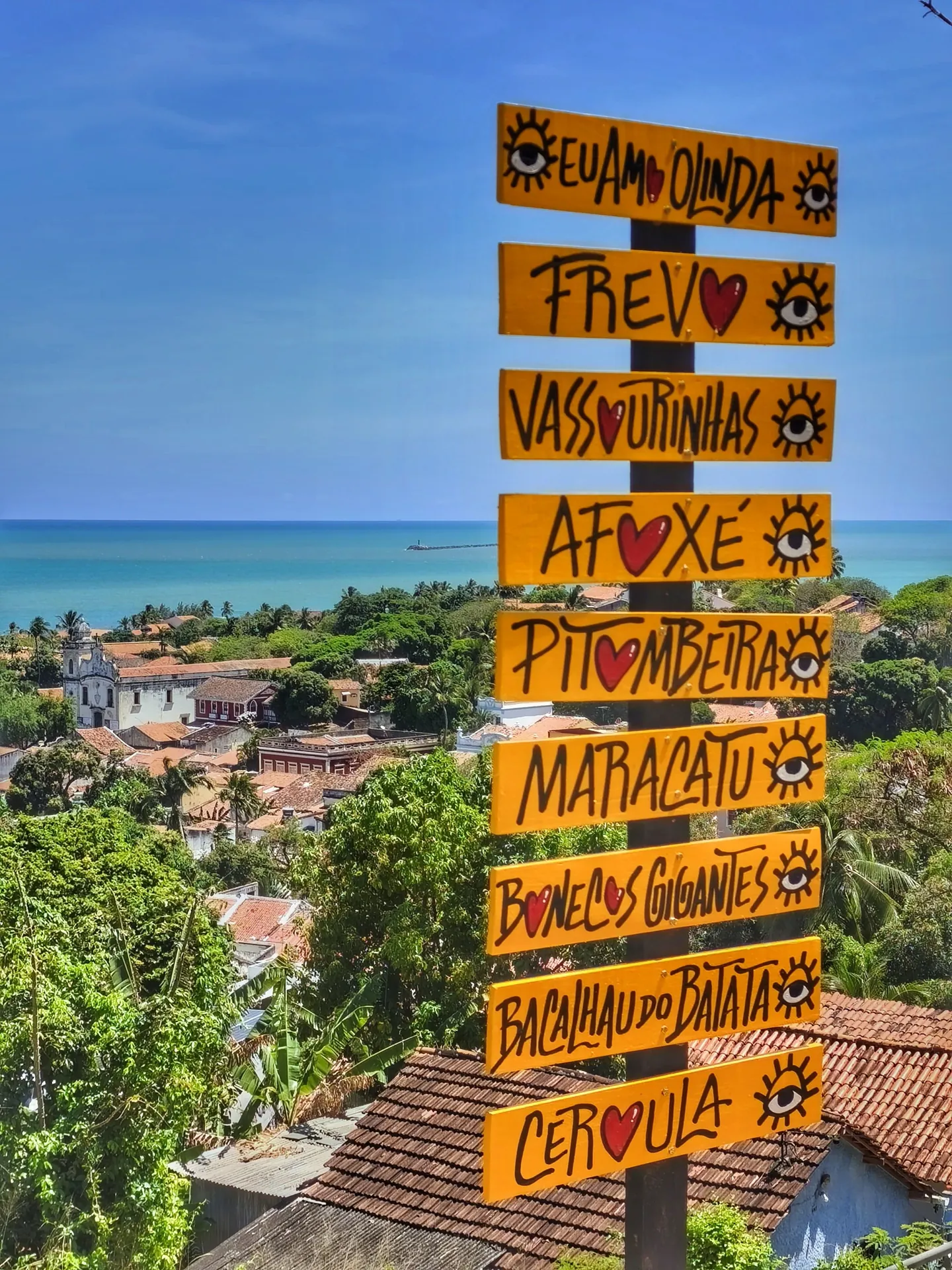 Placas de madeira pintadas à mão com nomes de blocos e ritmos de Olinda, com o casario e o mar ao fundo