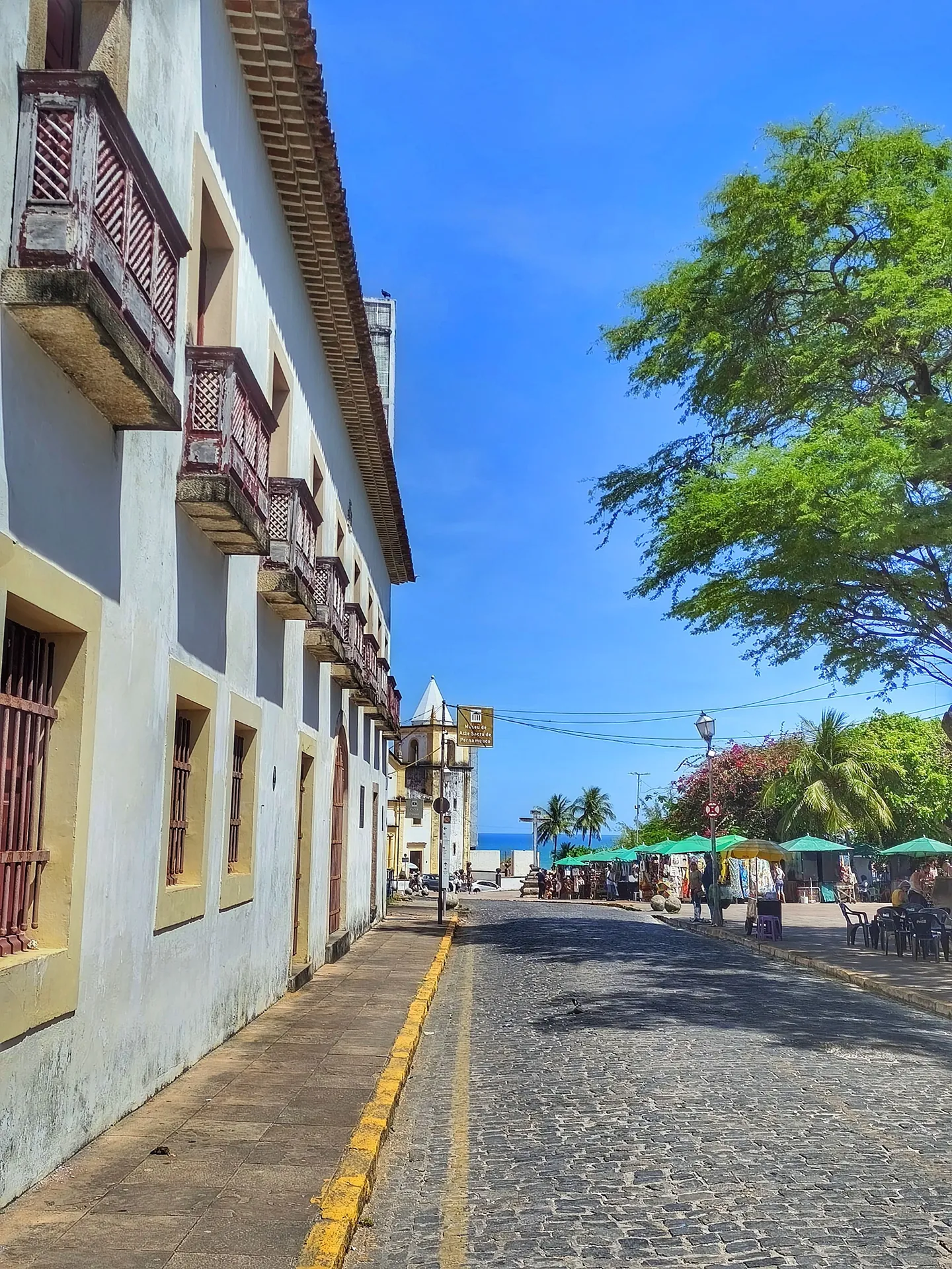 Rua de pedra no Sítio Histórico de Olinda, com casario branco e amarelo de sacadas e o mar ao fundo